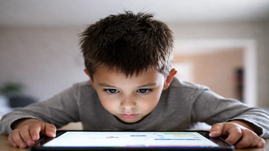 “Niño en edad escolar muy cerca de una tablet, con postura inclinada y mirada fija, señal de esfuerzo visual durante el uso prolongado de pantallas.”