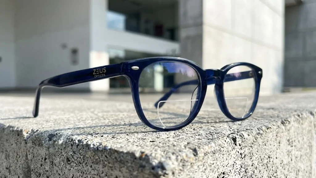 "Primer plano de las gafas bifocales premium de ZEUS ÓPTICOS en azul marino. El sol ilumina el diseño, destacando la precisión de la 'oblea' o segmento bifocal sobre una superficie de concreto arquitectónico moderno."
