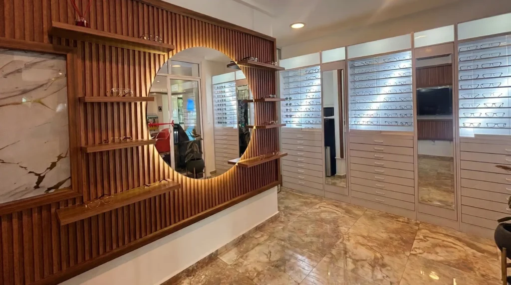 Interior moderno y elegante de una optica cerca de ti, destacando un panel de madera acanalada con un espejo redondo retroiluminado, estantes flotantes exhibiendo armazones y amplias vitrinas blancas con múltiples pares de lentes.