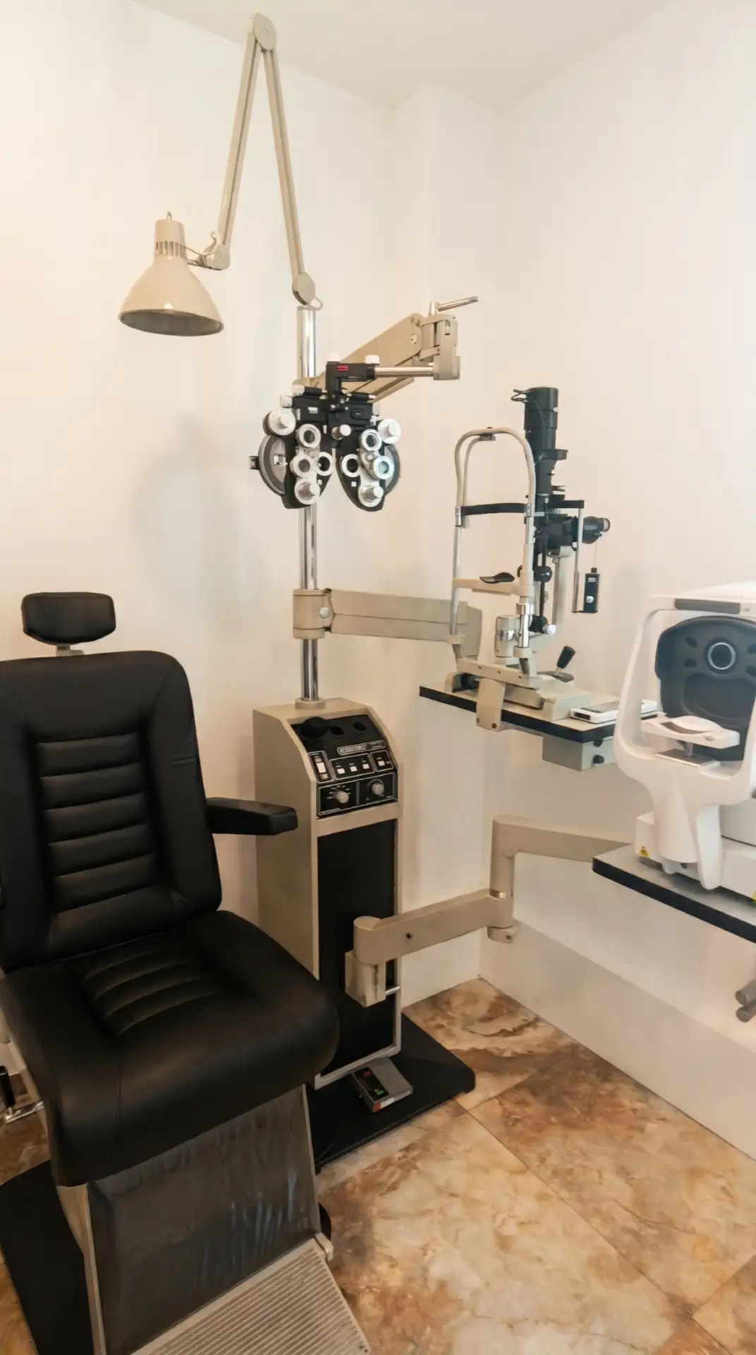 Consultorio optométrico equipado con una silla de exploración de vinipiel negra, una unidad oftalmológica que sostiene un foróptero, una lámpara de hendidura y un autorrefractor sobre un piso de mármol.